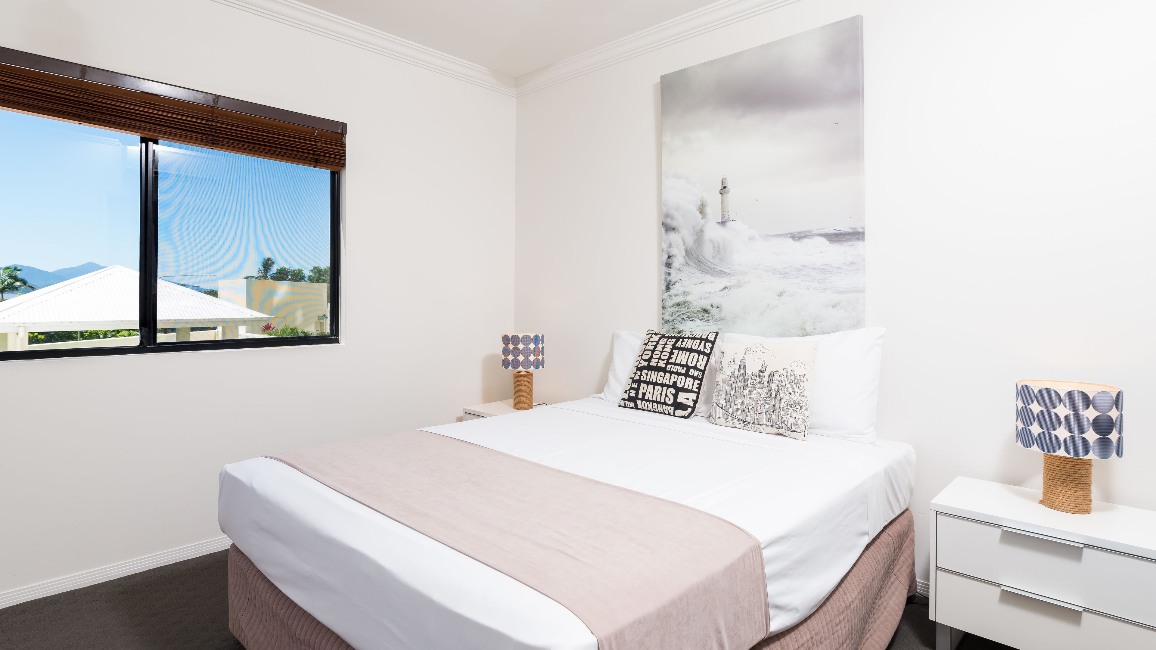 Un dormitorio ordenado con cama, mesitas de noche, ventana con persianas y un cuadro de un paisaje urbano en la pared.