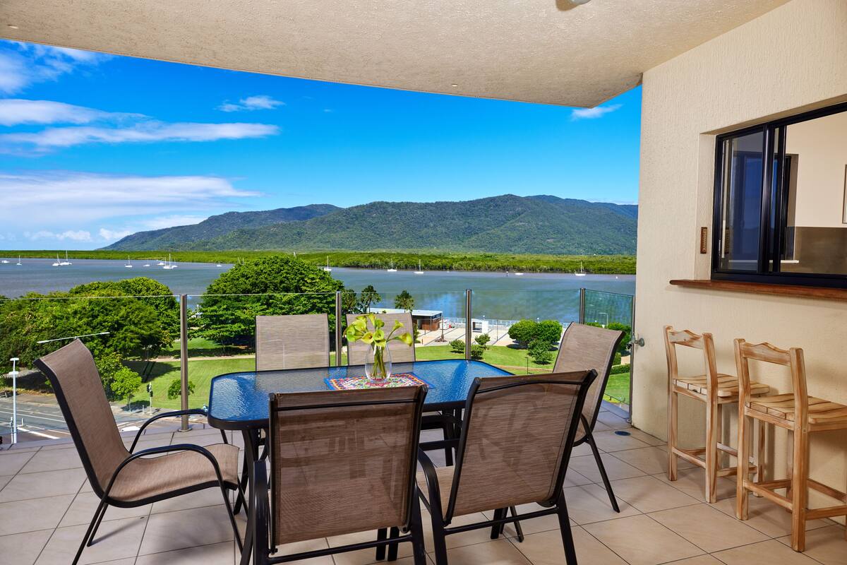 A balcony with a table and chairs, overlooking a lake with boats and mountains.