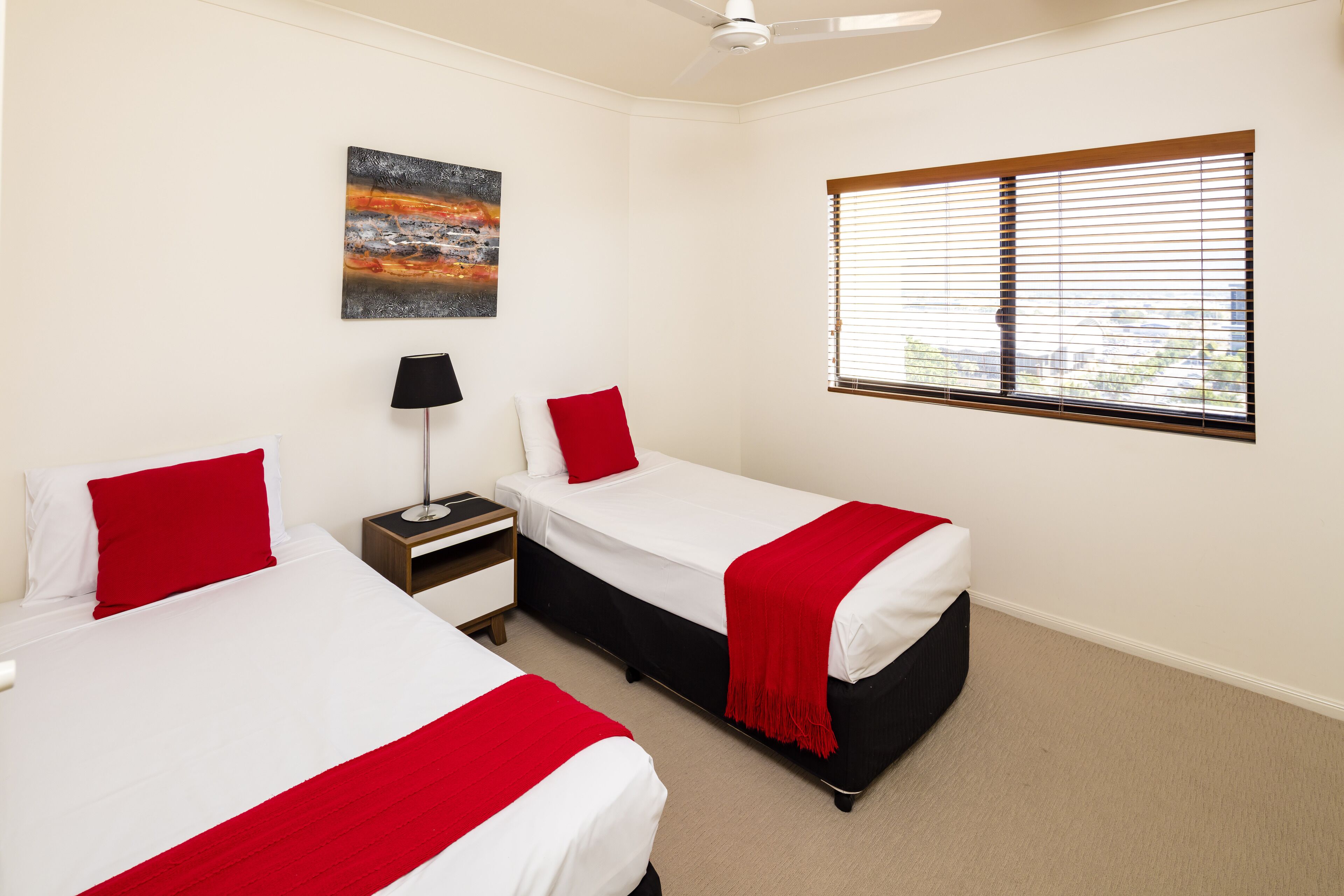 A hotel room with two beds, a nightstand, a lamp, a painting, and a window with blinds.