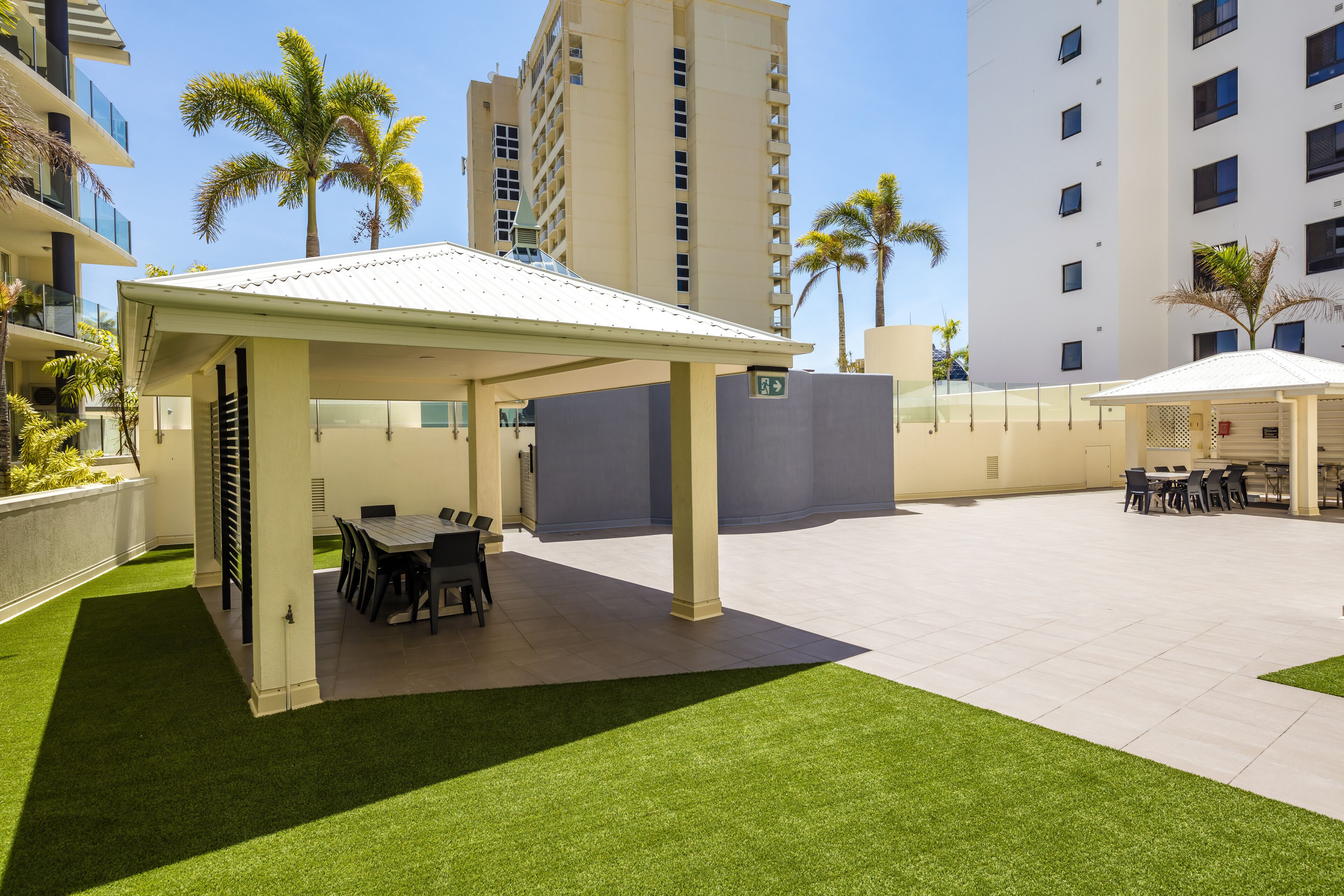 A covered outdoor area with a dining table and chairs, surrounded by artificial grass and palm trees.