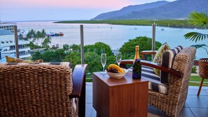 A wicker sofa and table with a bottle of champagne, a bowl of fruit, and two glasses on a balcony overlooking a marina and mountains. - Jack & Newell Holiday Apartments (Cairns)