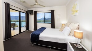 A hotel room with a large bed, a ceiling fan, a view of a mountain and lake, and a balcony with a chair. - Jack & Newell Holiday Apartments (Cairns)