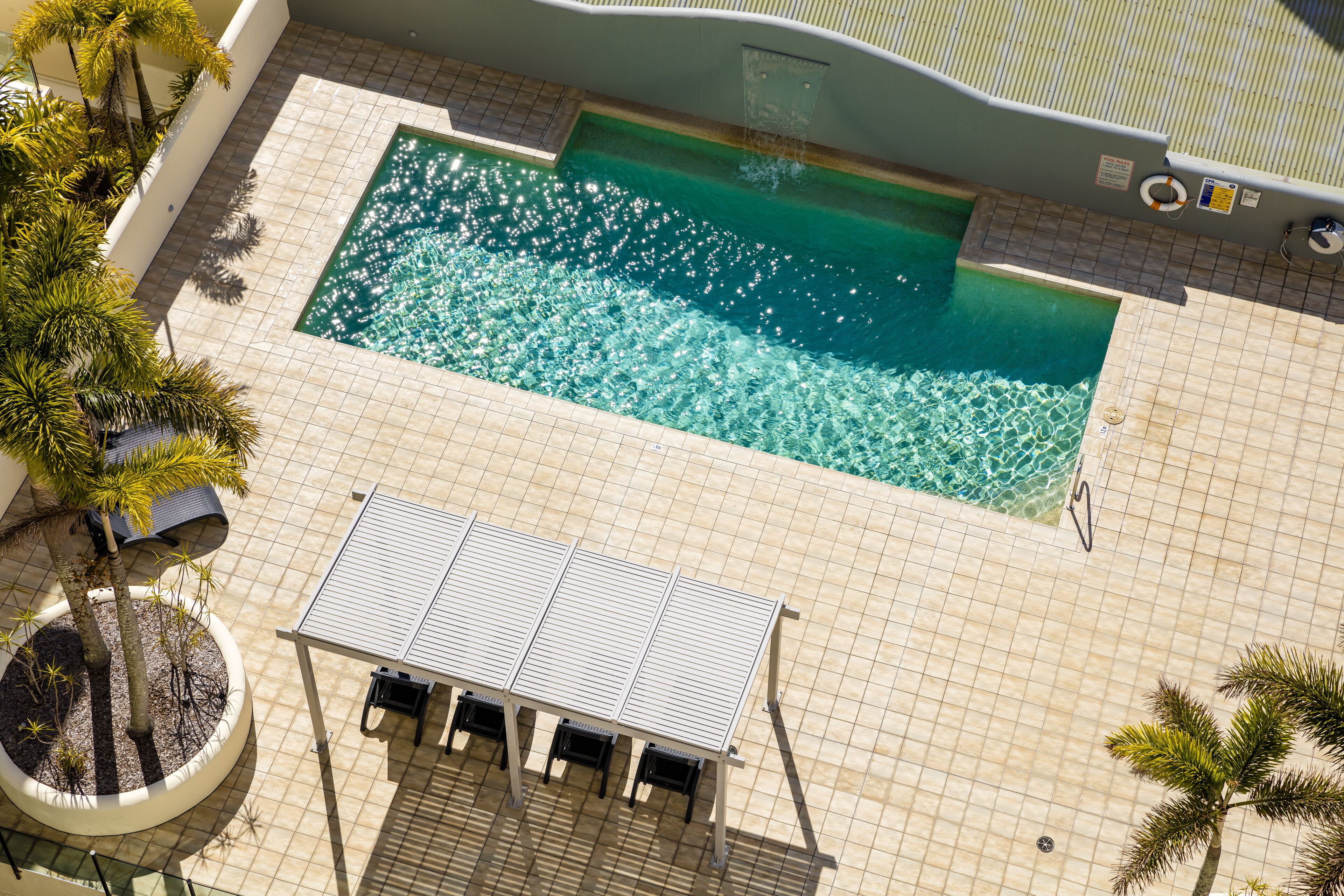 Vista aérea de uma piscina com piso em azulejos, um espaço coberto para sentar e palmeiras ao redor.