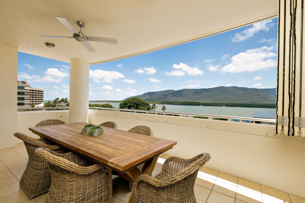 A balcony with a wooden table and wicker chairs, overlooking a lake and mountains.