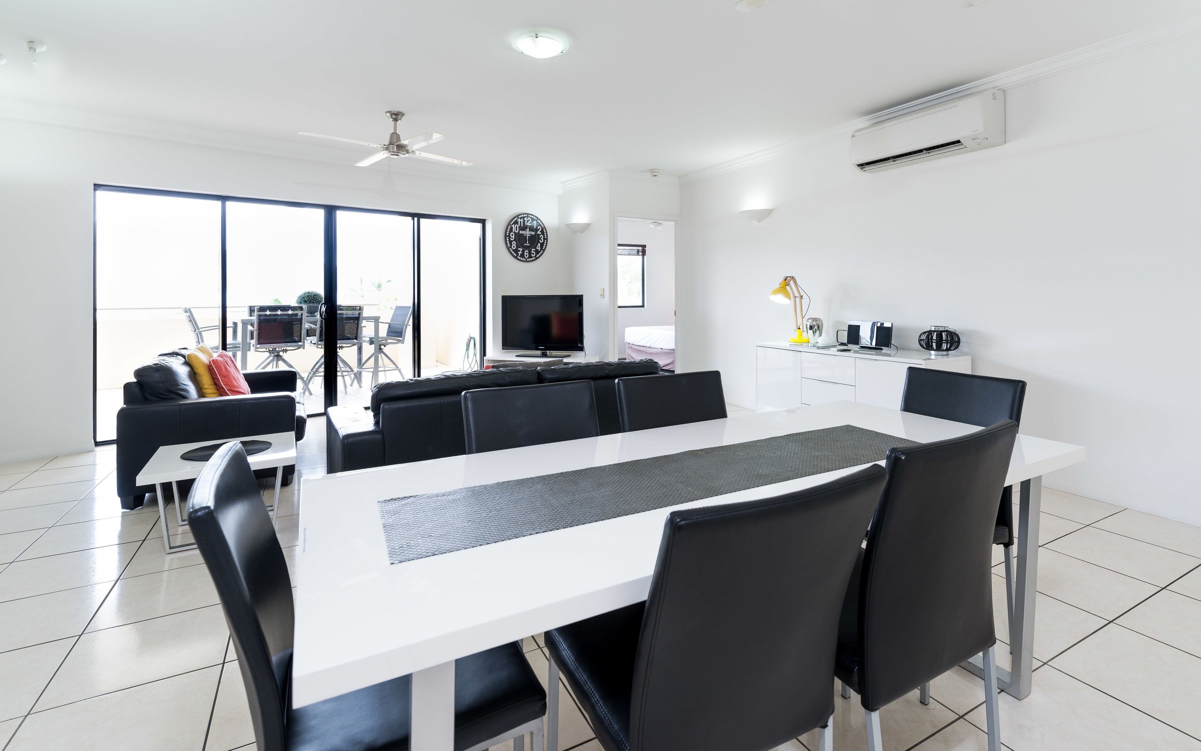 A modern living room with a large white dining table, black chairs, a sofa, and a TV.