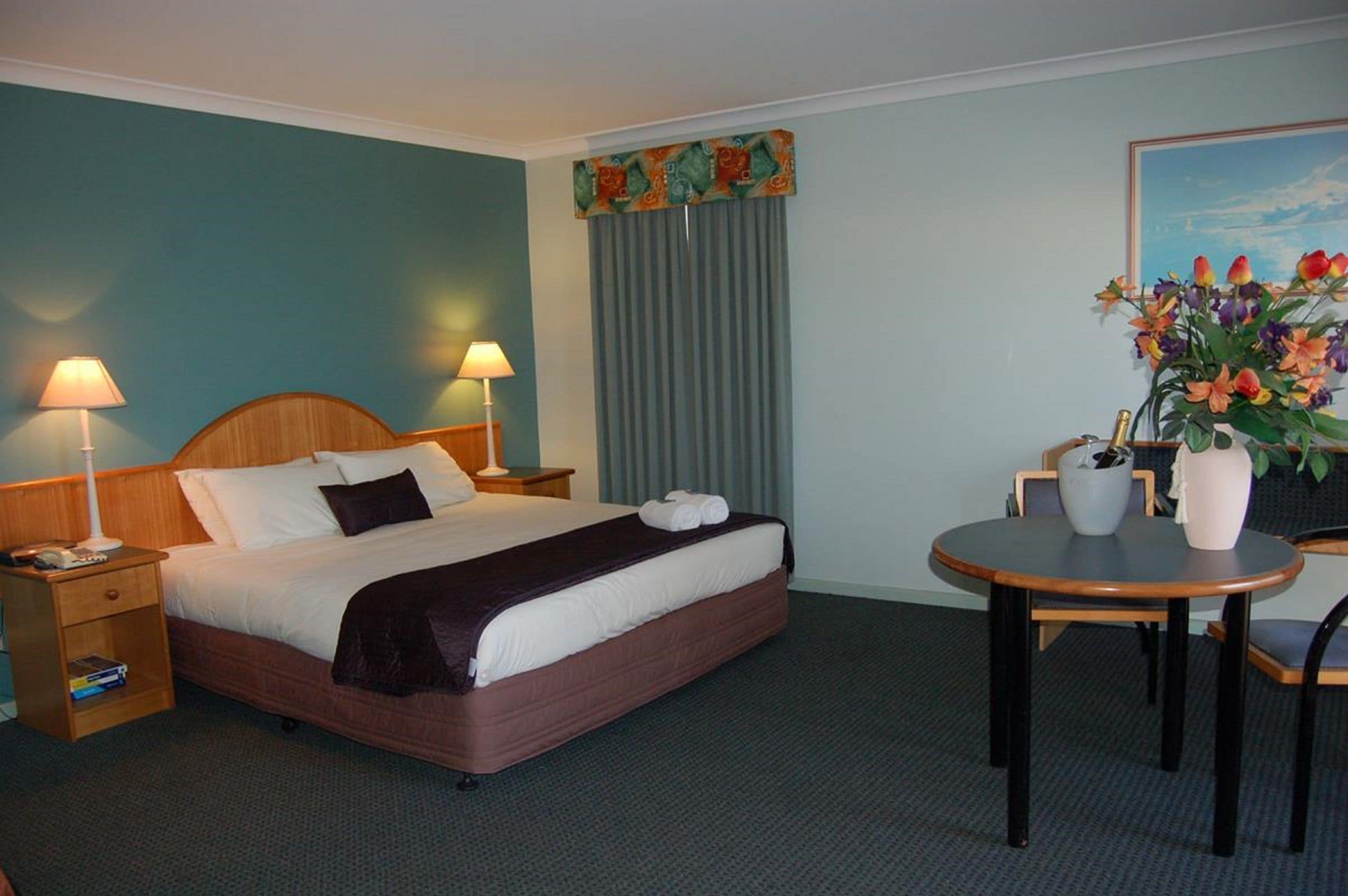 A hotel room with a bed, two bedside lamps, a small table with chairs, a vase with flowers, and a painting on the wall.