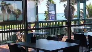 An outdoor dining area with tables and chairs, overlooking a beach with palm trees and a view of the ocean. - Heritage Resort Shark Bay (Denham)