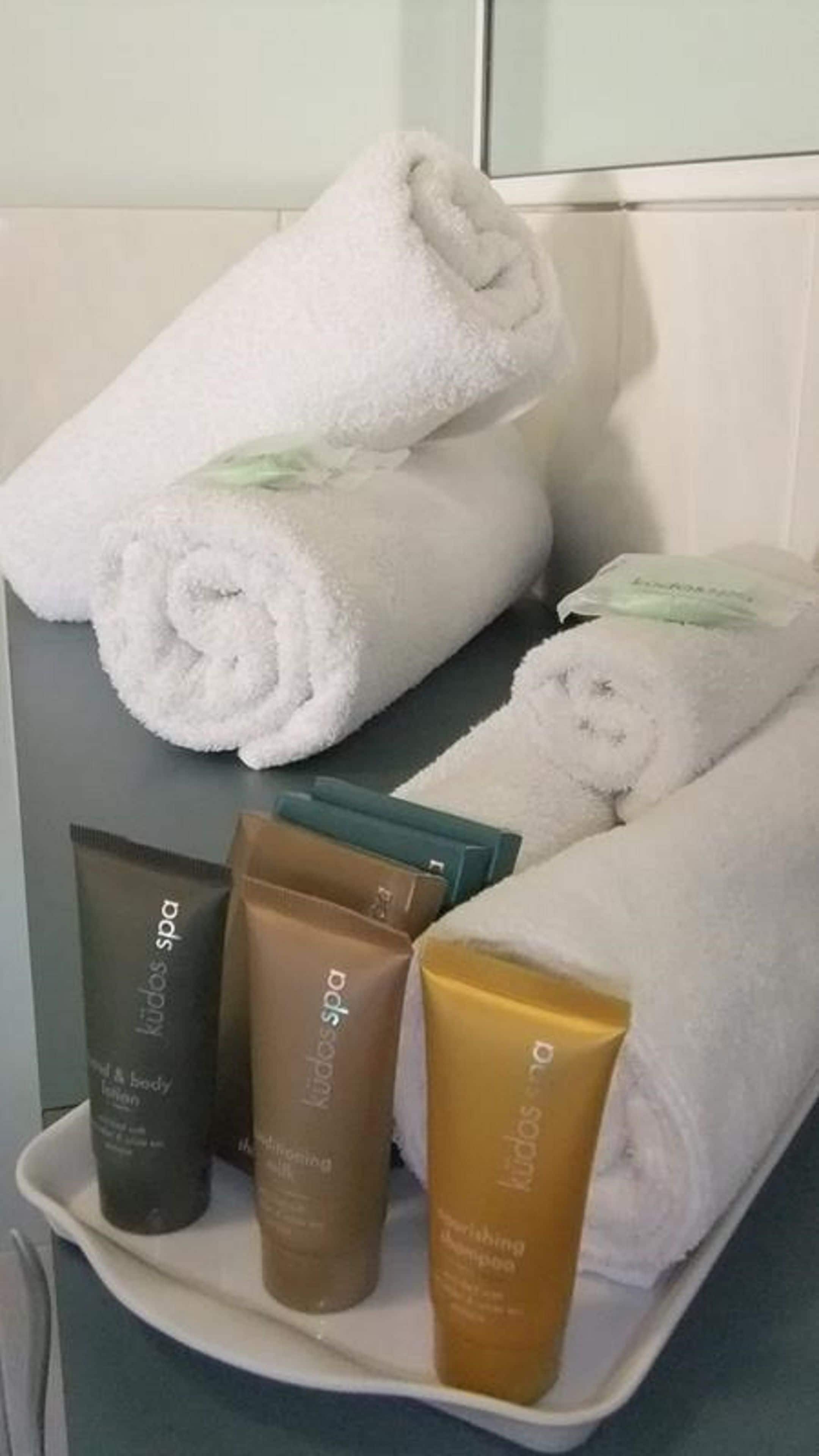 Un baño con toallas blancas ordenadas, una bandeja con tres productos de 'Kudos Spa' y un espejo.