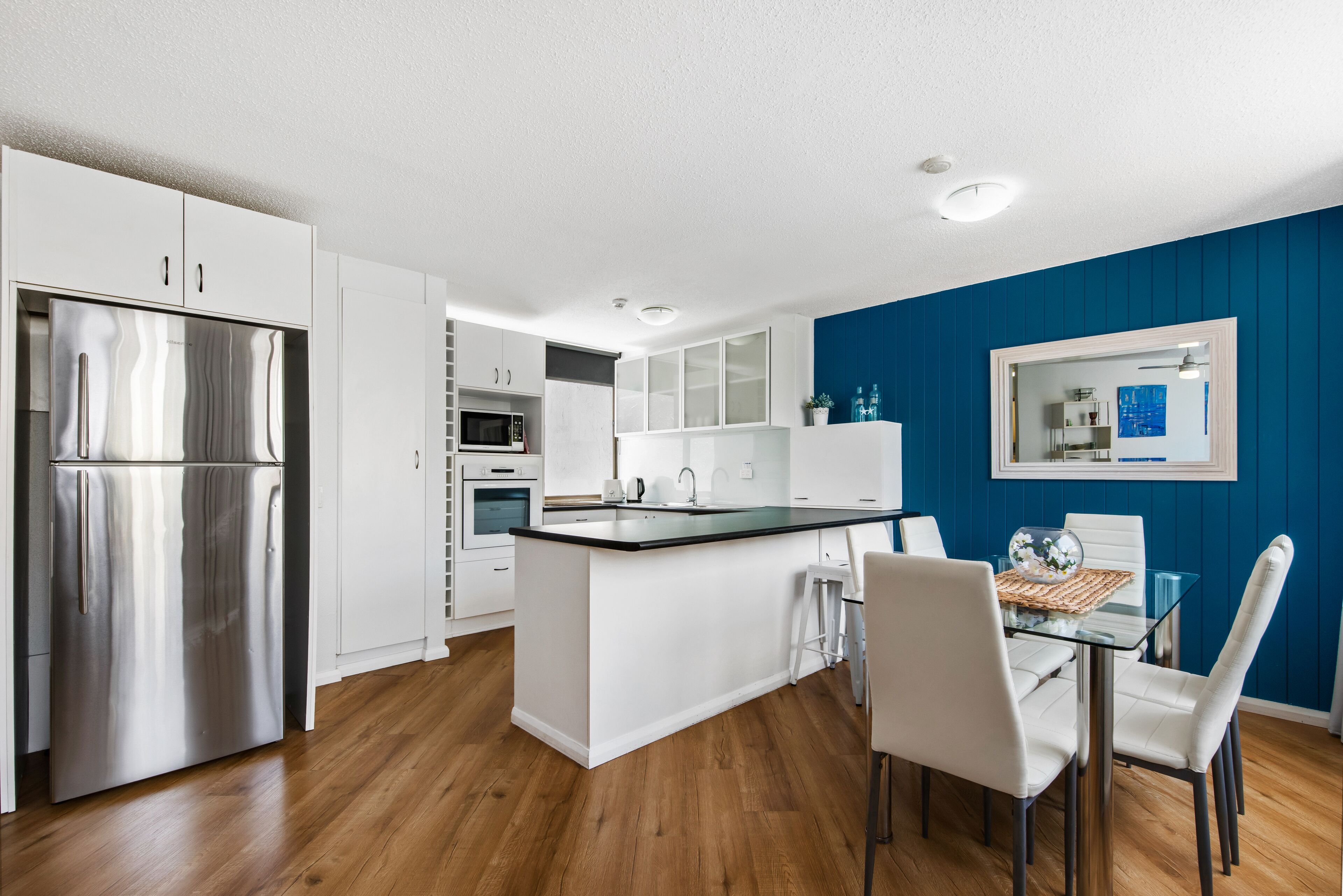 A modern kitchen with white cabinetry, a stainless steel refrigerator, a central island, and a dining area with white chairs.