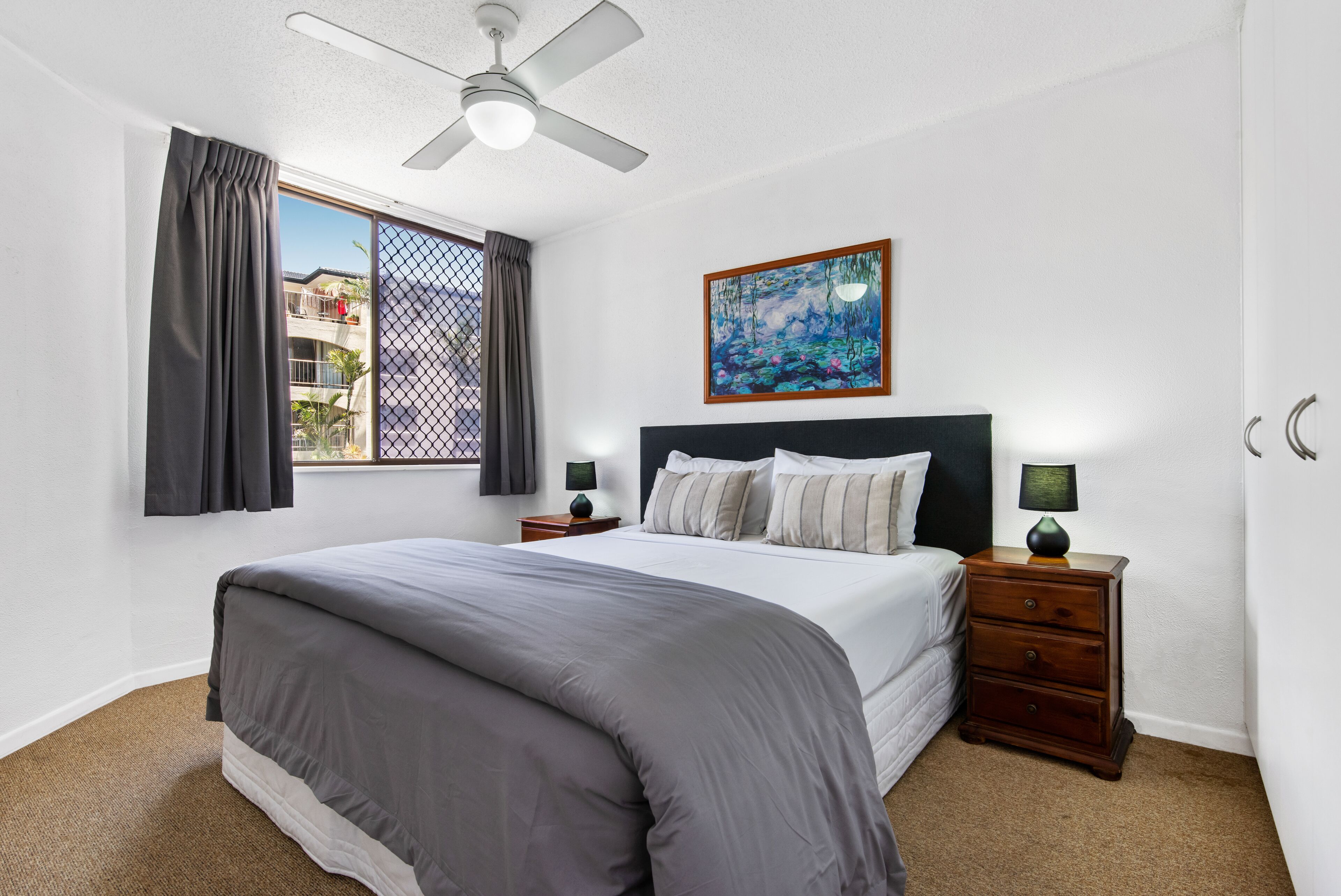 A bedroom with a bed, a ceiling fan, a window with curtains, a painting, and a nightstand with a lamp.