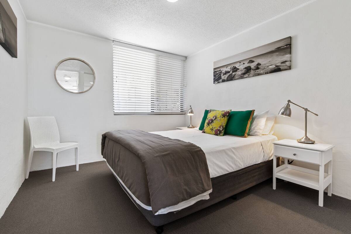 A bedroom with a bed, a bedside table, a lamp, a chair, a window with blinds, a round mirror, and a wall-mounted picture.