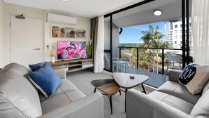 A modern living room with a sofa, coffee table, and TV. There is a balcony with a view of palm trees and buildings. - Mylos Holiday Apartments (Maroochydore)