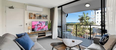 A modern living room with a sofa, coffee table, and TV. There is a balcony with a view of palm trees and buildings.