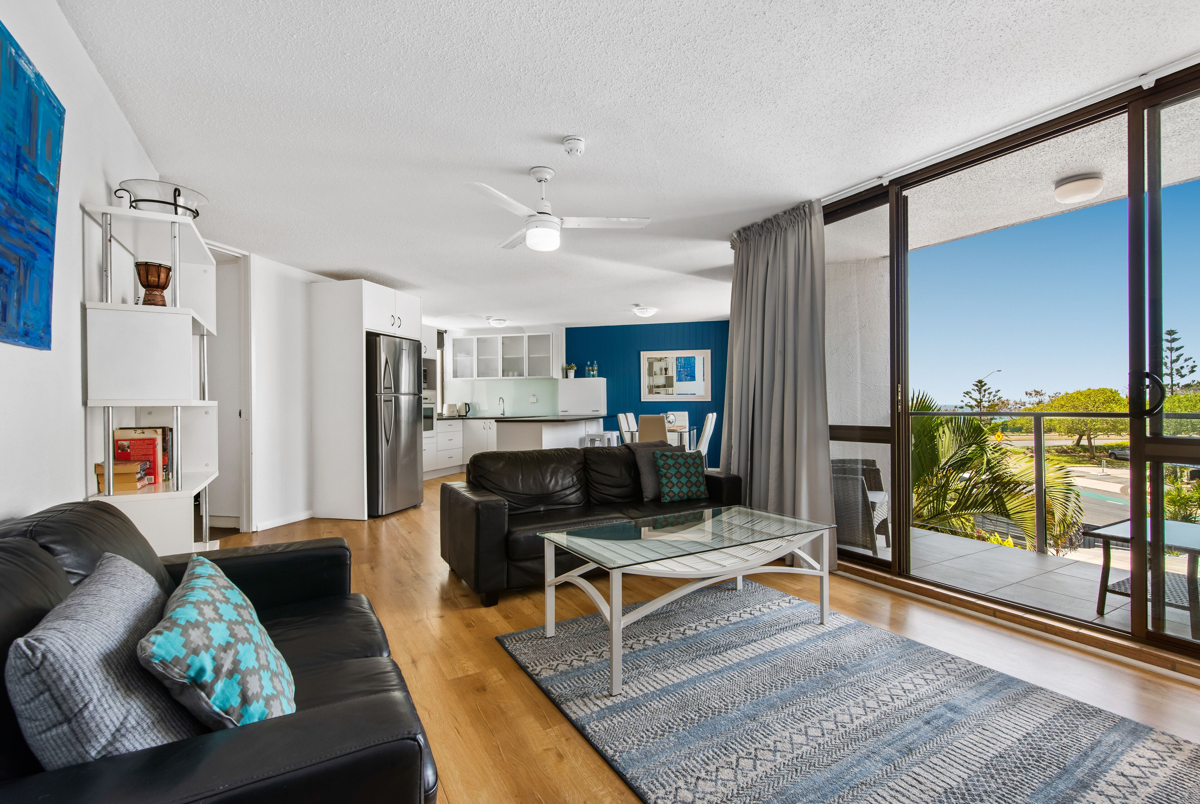 A modern living room with a glass coffee table, a black leather sofa, and a view of the outdoor balcony.