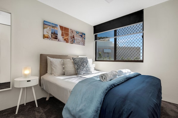 A bedroom with a bed, a bedside table with a lamp, a window with a view, and a wall with a picture.