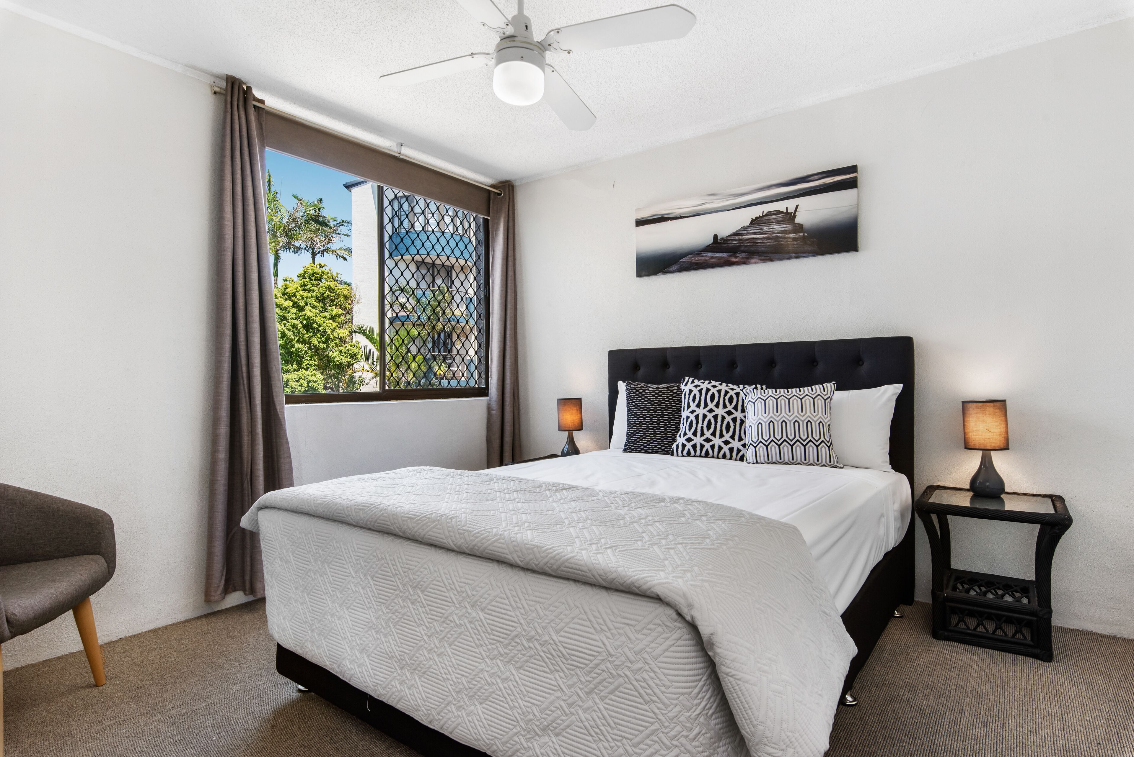 A bedroom with a bed, two bedside lamps, a ceiling fan, a window with curtains, and a wall-mounted picture.