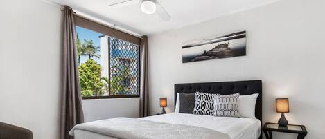 A bedroom with a bed, two bedside lamps, a ceiling fan, a window with curtains, and a wall-mounted picture.