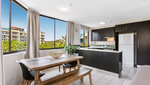 A modern kitchen with dark cabinets, a white refrigerator, and a large window overlooking a green landscape. - Mylos Holiday Apartments (Maroochydore)