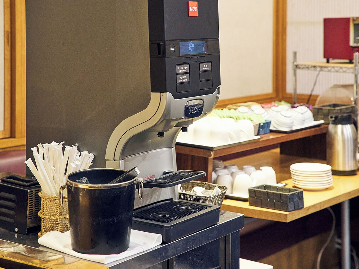 Una cafetera con pantalla digital, un cesto de pajitas, un termo y una pila de platos sobre una encimera.