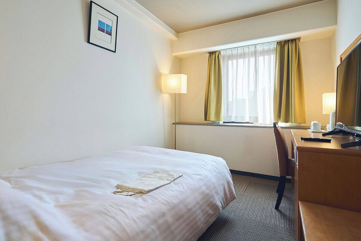 A hotel room with a bed, a desk, a chair, a lamp, and a window with curtains.