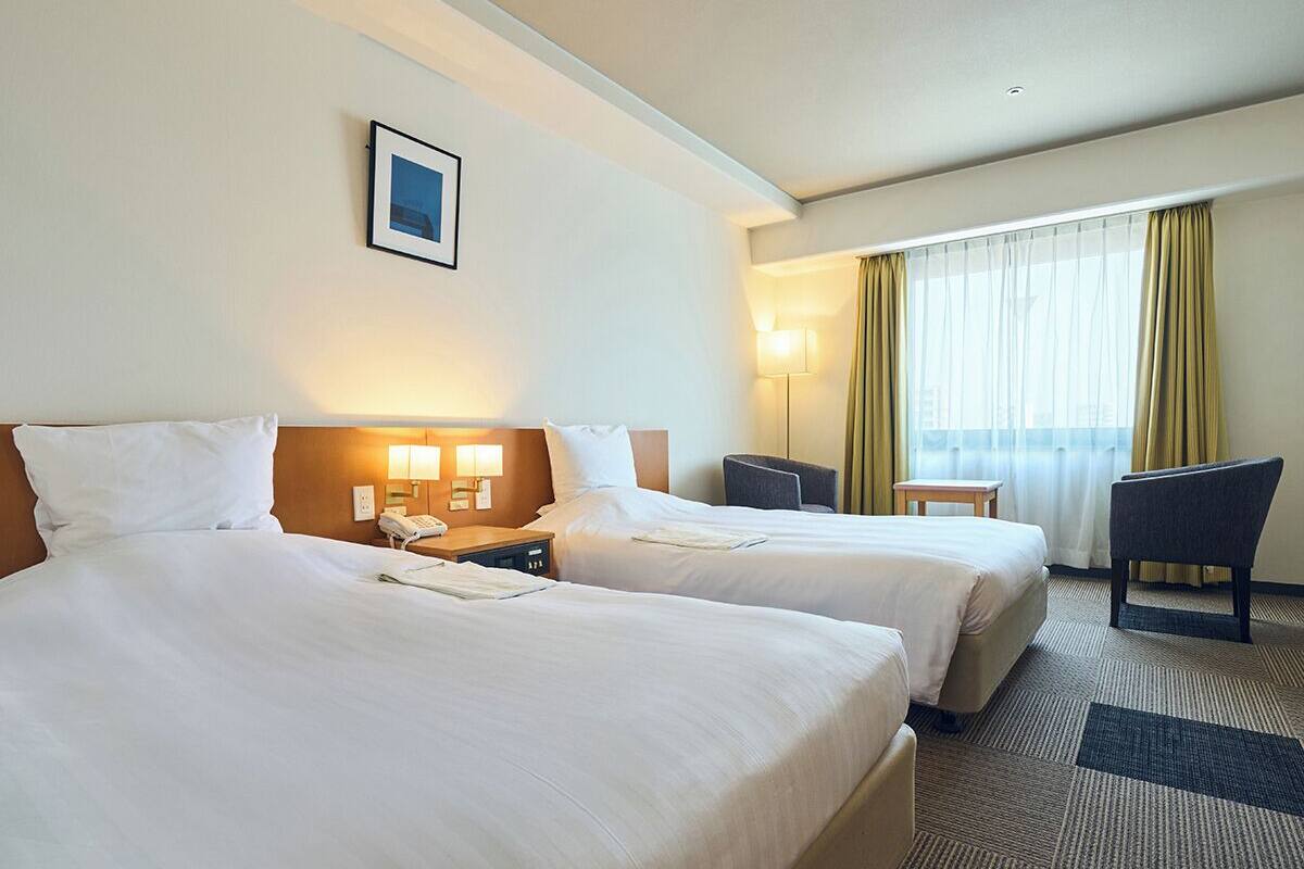 A hotel room with two beds, a desk, a chair, a lamp, and a window with curtains.