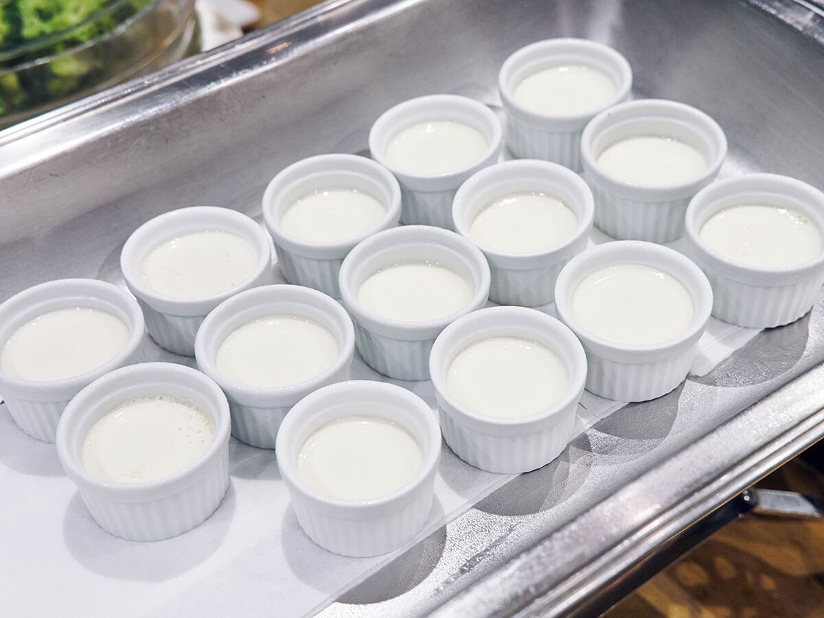 A tray of white ramekins with a creamy substance, likely custard or pudding, arranged neatly on a metallic surface.