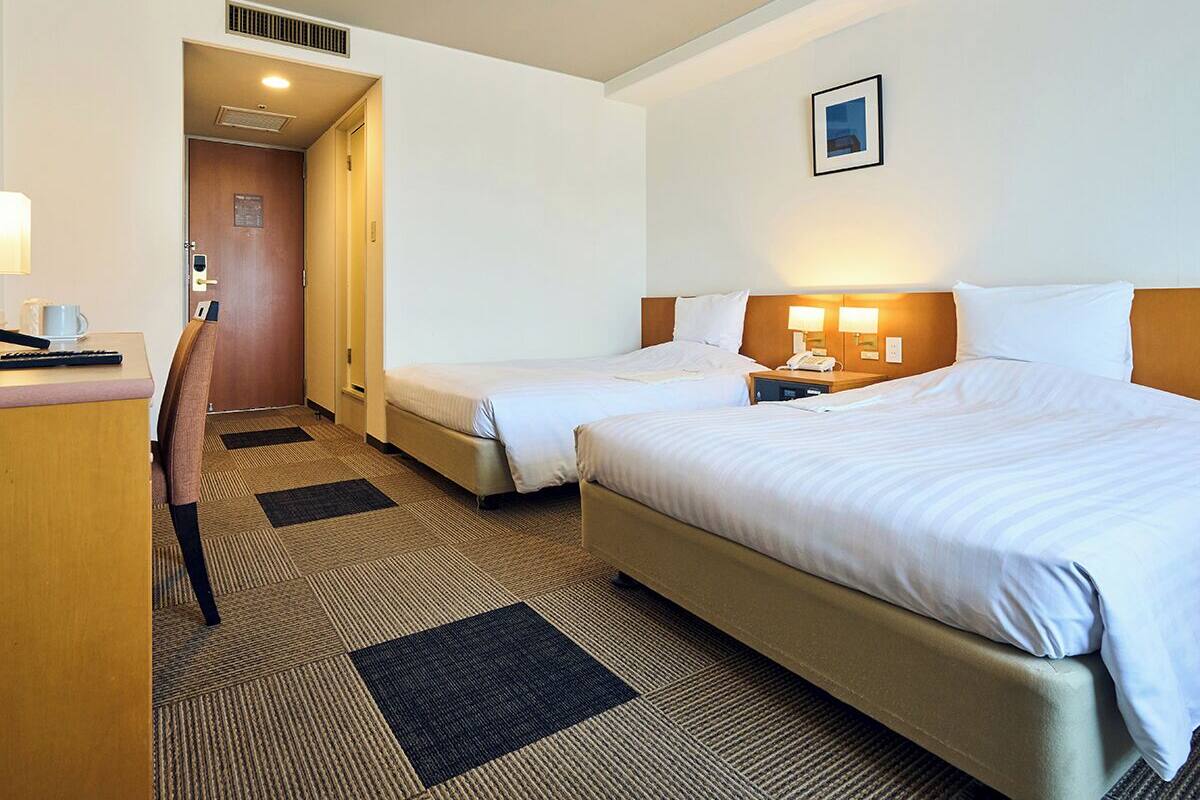 A hotel room with two beds, a desk, and a chair. There is a door leading to a hallway.