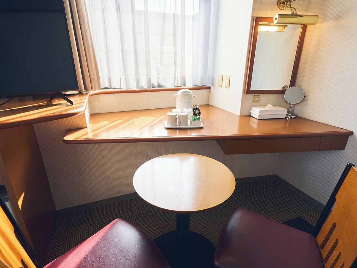 A hotel room with a wooden desk, a round table, a chair, a TV, a window with sheer curtains, a bedside table with a lamp, and a mirror.