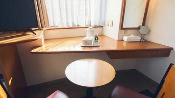 A hotel room with a wooden desk, a round table, a chair, a TV, a window with sheer curtains, a bedside table with a lamp, and a mirror.