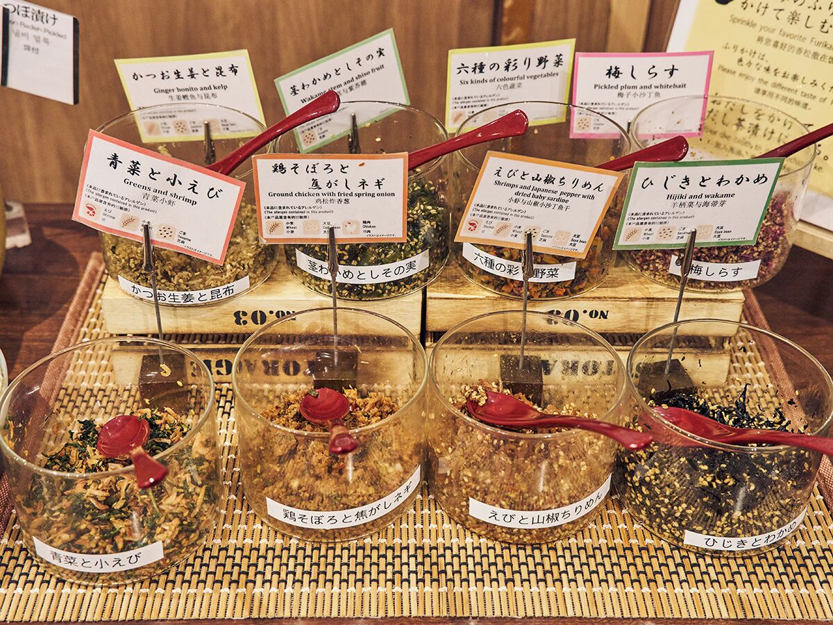 Una exhibición de diversos alimentos encurtidos y marinados en recipientes de vidrio con etiquetas, colocados sobre un tapete tejido.