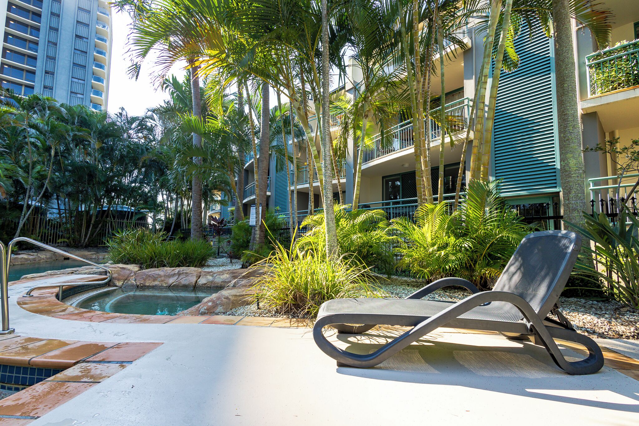 A pool area with lounge chairs, palm trees, and a building with balconies.