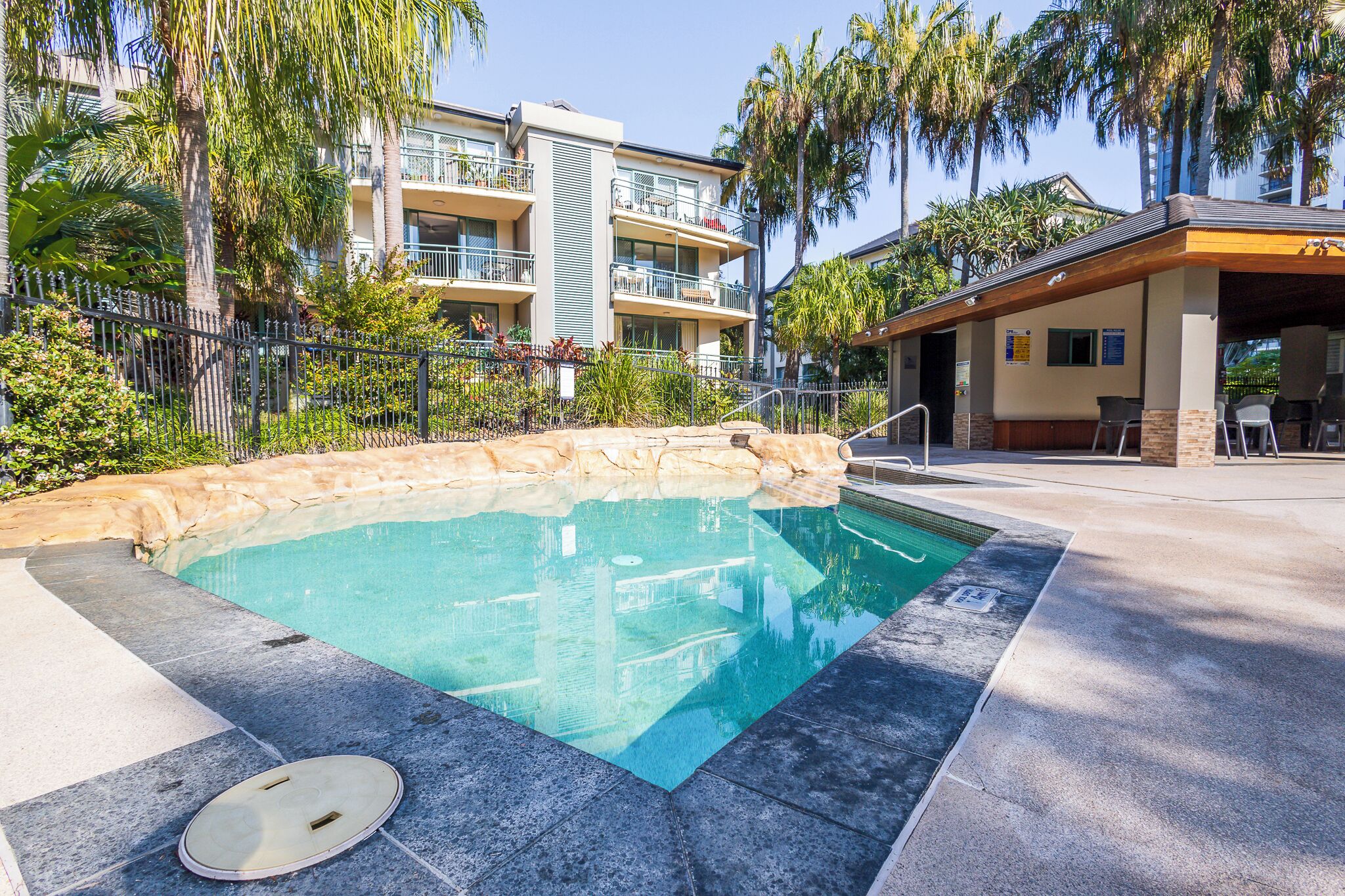 Una piscina con borde de piedra, rodeada por un área pavimentada y una valla.