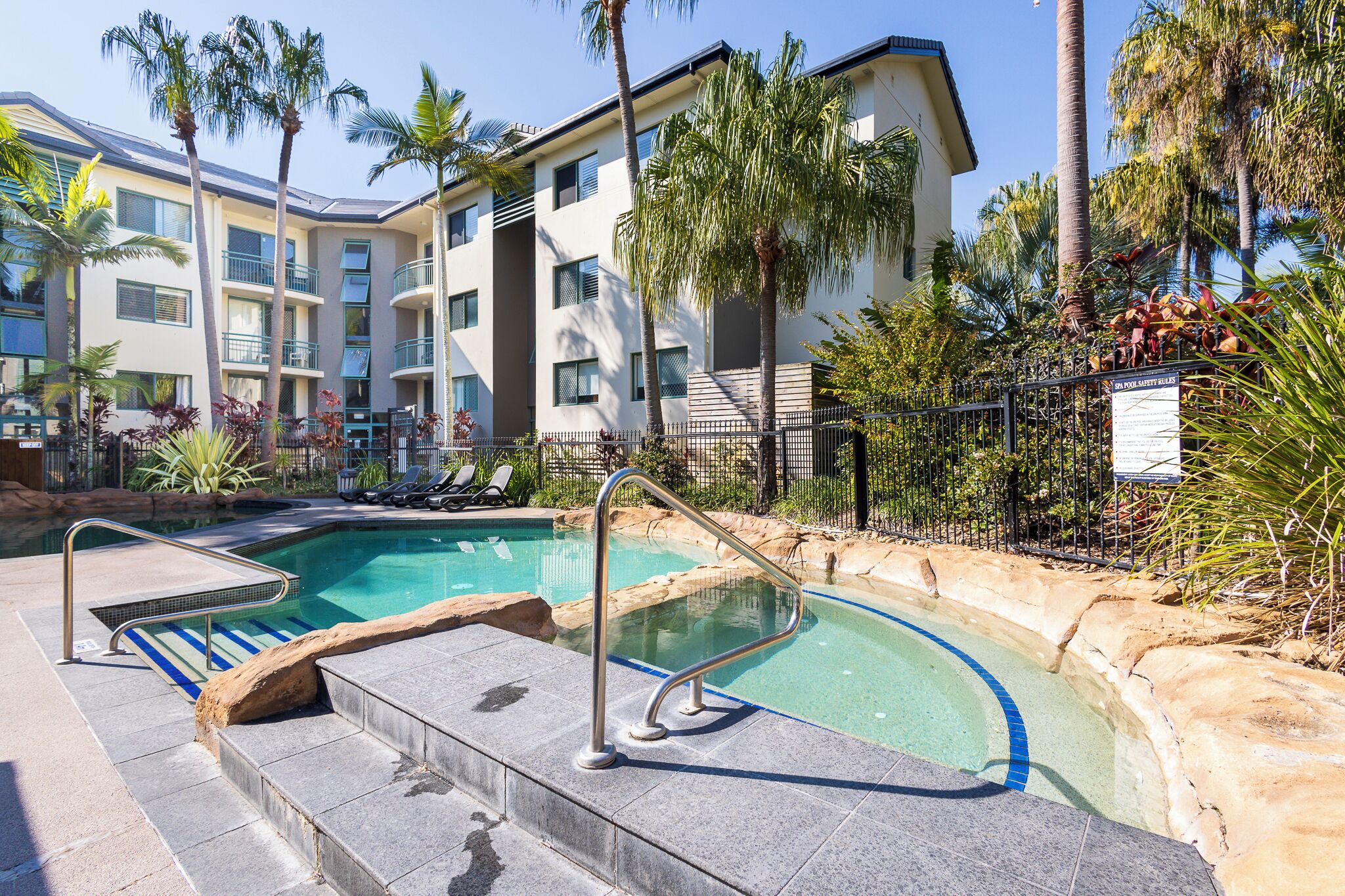 A swimming pool with a stone border, palm trees, and a multi-story building with balconies.