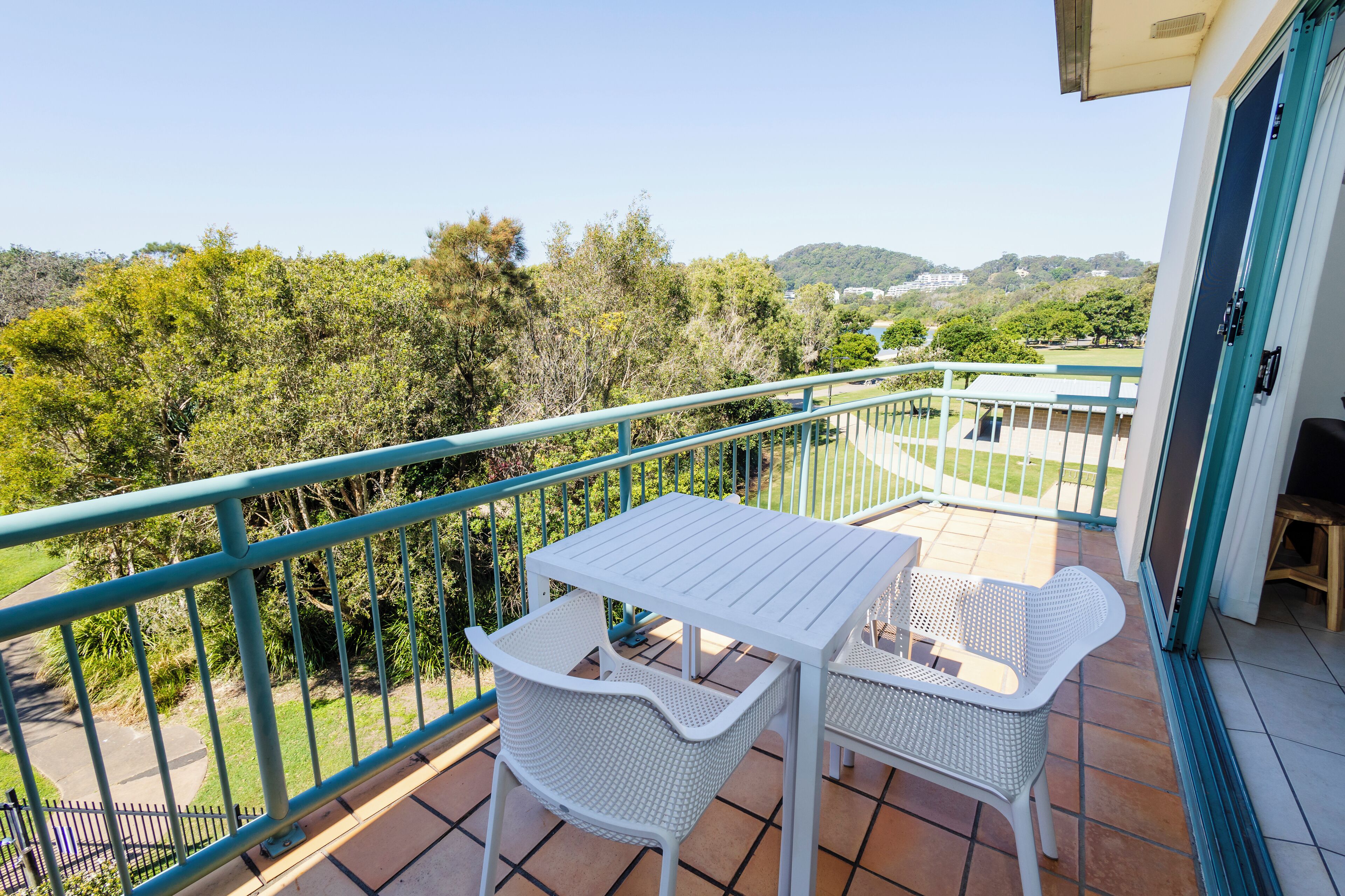 A balcony with a white table and two white chairs, offering a view of a green landscape and hills.