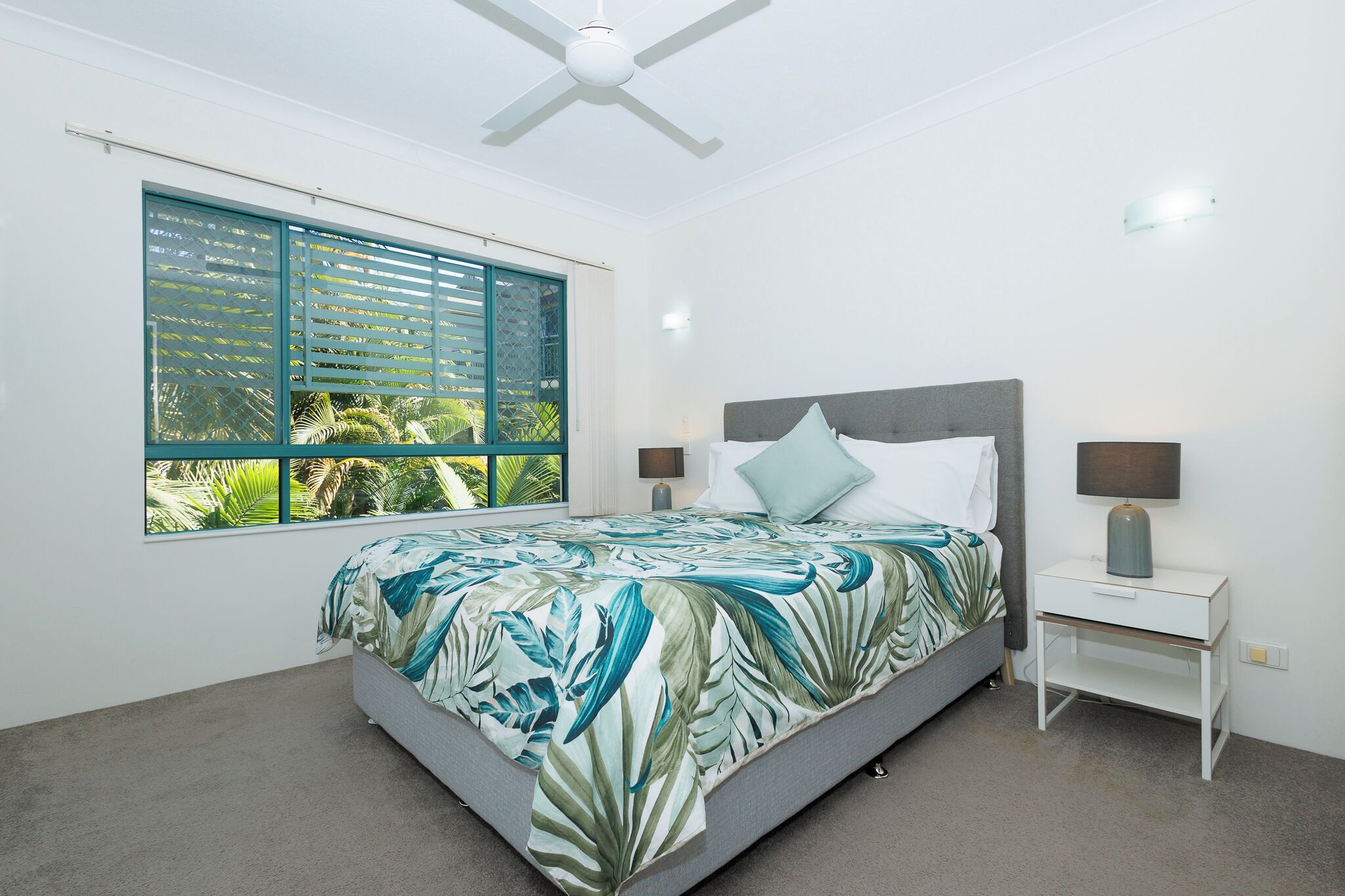 A bedroom with a bed, a nightstand, a ceiling fan, a window with blinds, and a view of greenery outside.