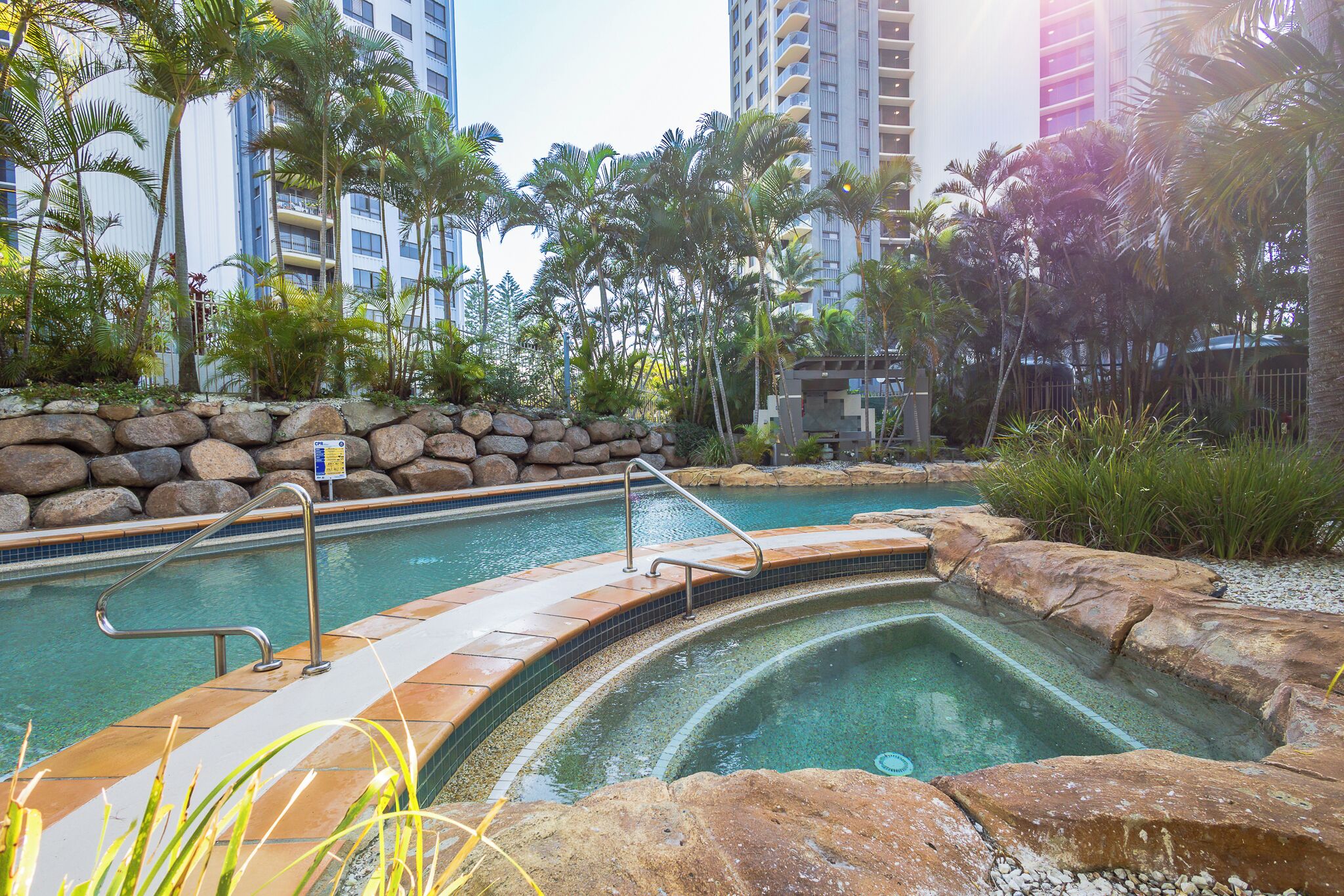 A swimming pool surrounded by rocks and palm trees, with a stone wall and metal railings.