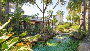 A tropical garden with a pond, palm trees, and a house with a balcony. - Currumbin Sands Holiday Apartments (Palm Beach)
