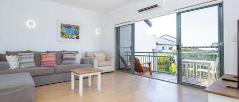 A spacious living room with a grey sofa, a wooden coffee table, and a balcony with a view of the sea.