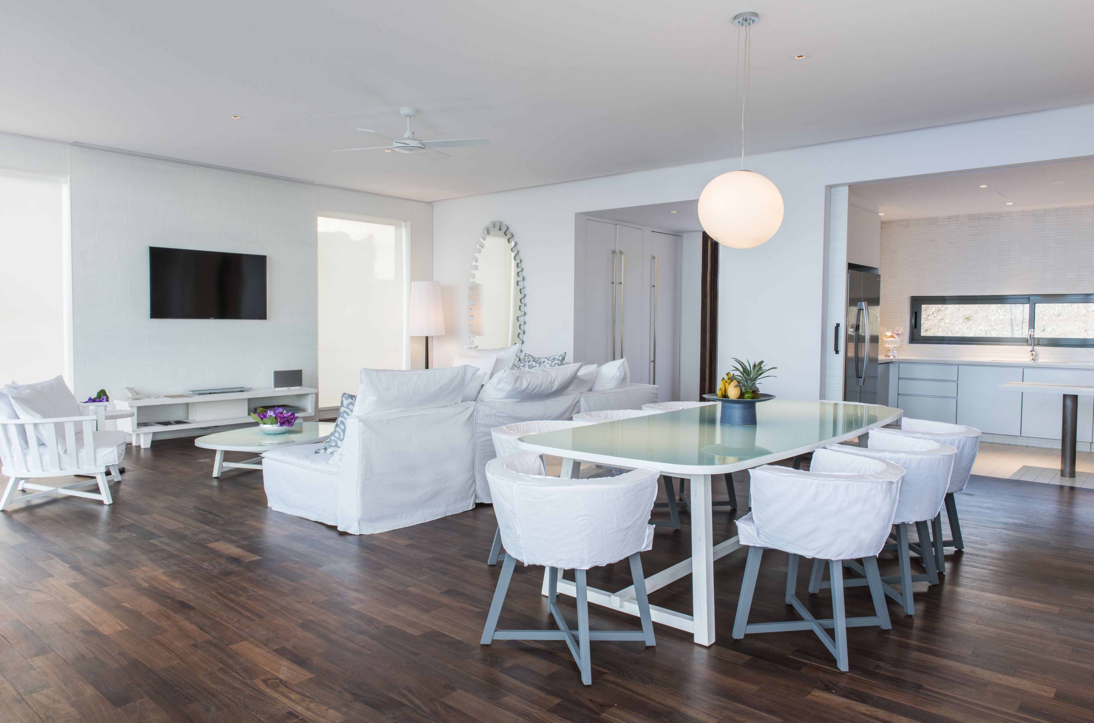 Una sala de estar moderna con una mesa de comedor de vidrio, sofás blancos y una cocina visible a través de un umbral abierto.