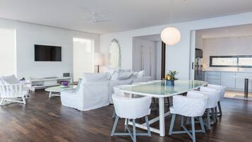 Una sala de estar moderna con una mesa de comedor de vidrio, sofás blancos y una cocina visible a través de un umbral abierto.