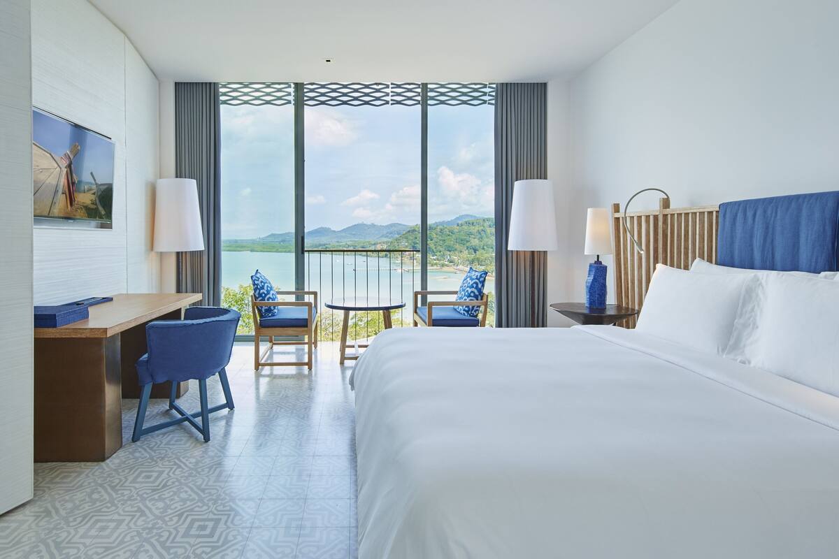 Een hotelkamer met een groot bed, twee stoelen, een bureau en een balkon met uitzicht op het landschap.