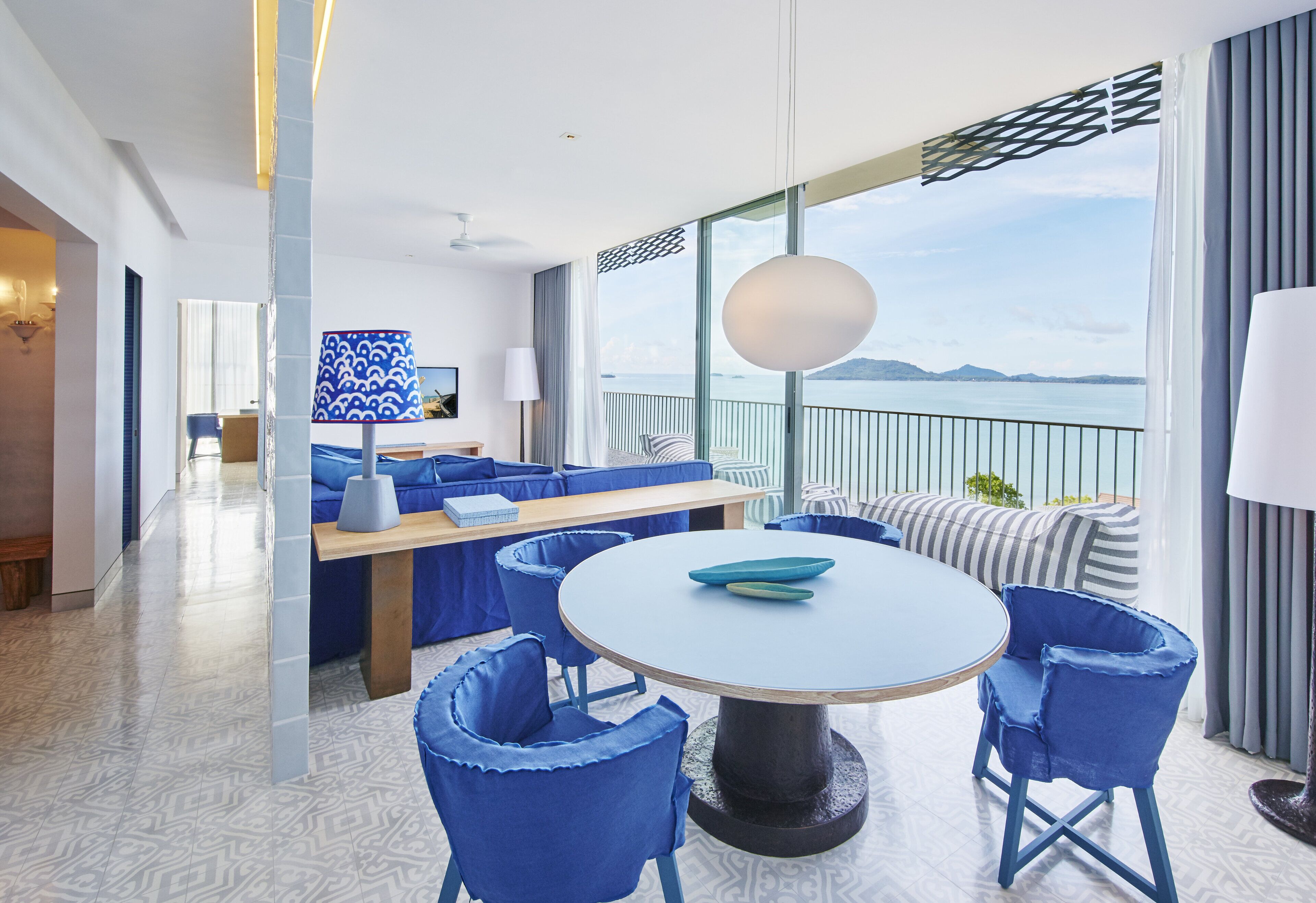 A modern hotel room with a round table, blue chairs, a sofa, and a balcony with a view of the sea.