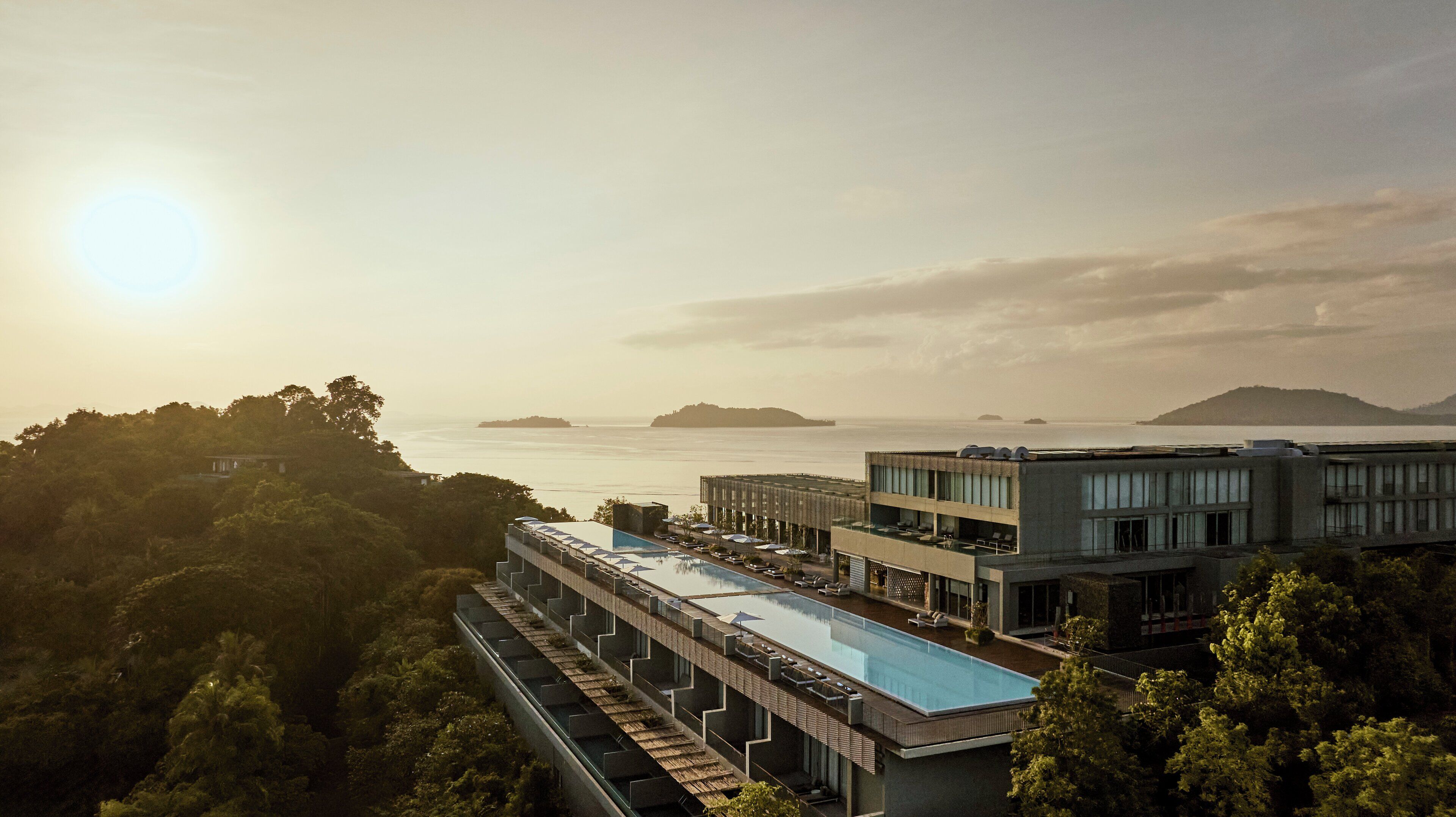 A modern hotel with a rooftop infinity pool, lush greenery, and a view of the sea and distant islands.