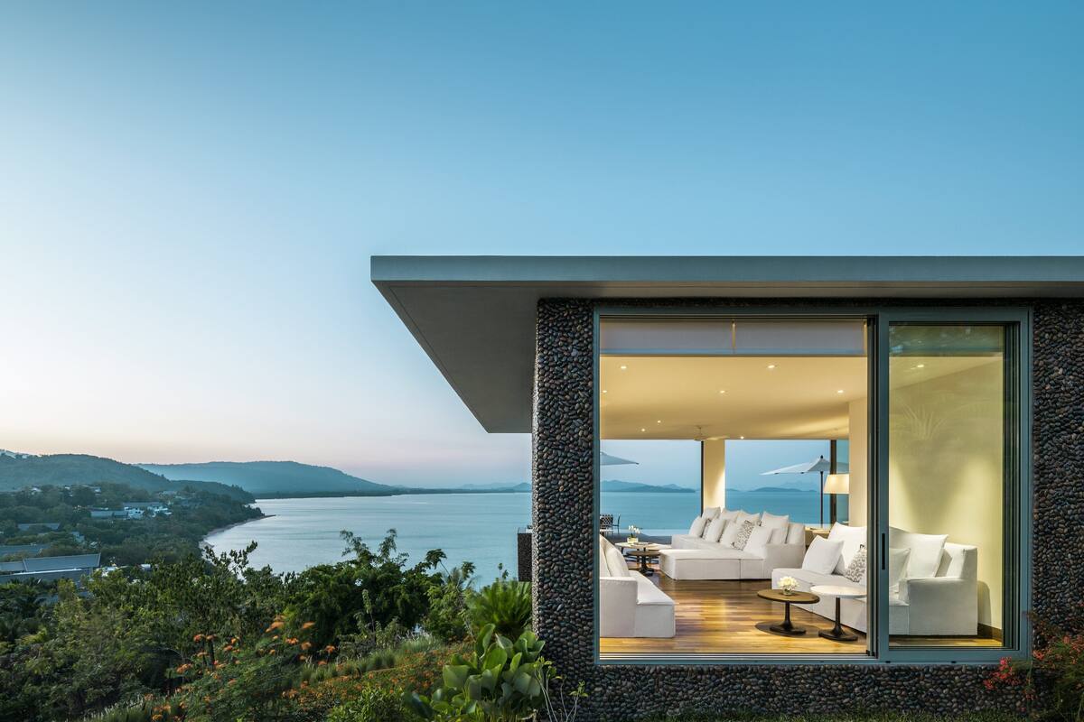 Een modern huis met een grote glazen deur die uitkomt op een woonkamer met een witte bank en een tafel, met uitzicht op een waterpartij en heuvels.