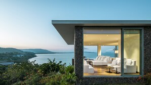 A modern house with a large glass door leading to a living area with a white sofa and a table, overlooking a body of water and hills.