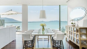 A dining area with a table set for six, a white sofa, a wooden cabinet, and a large window offering a view of the water and mountains.