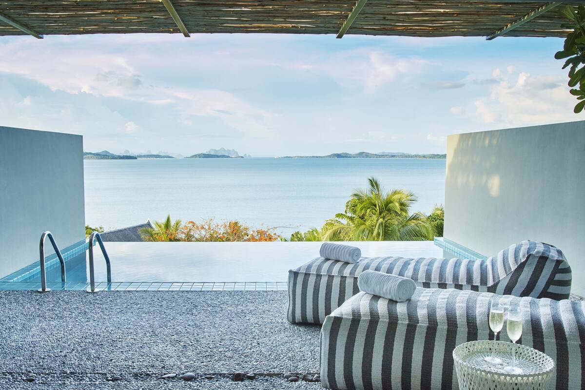 Een lounge aan het zwembad met uitzicht op zee, een overdekt gedeelte en een rieten tafel met glazen.