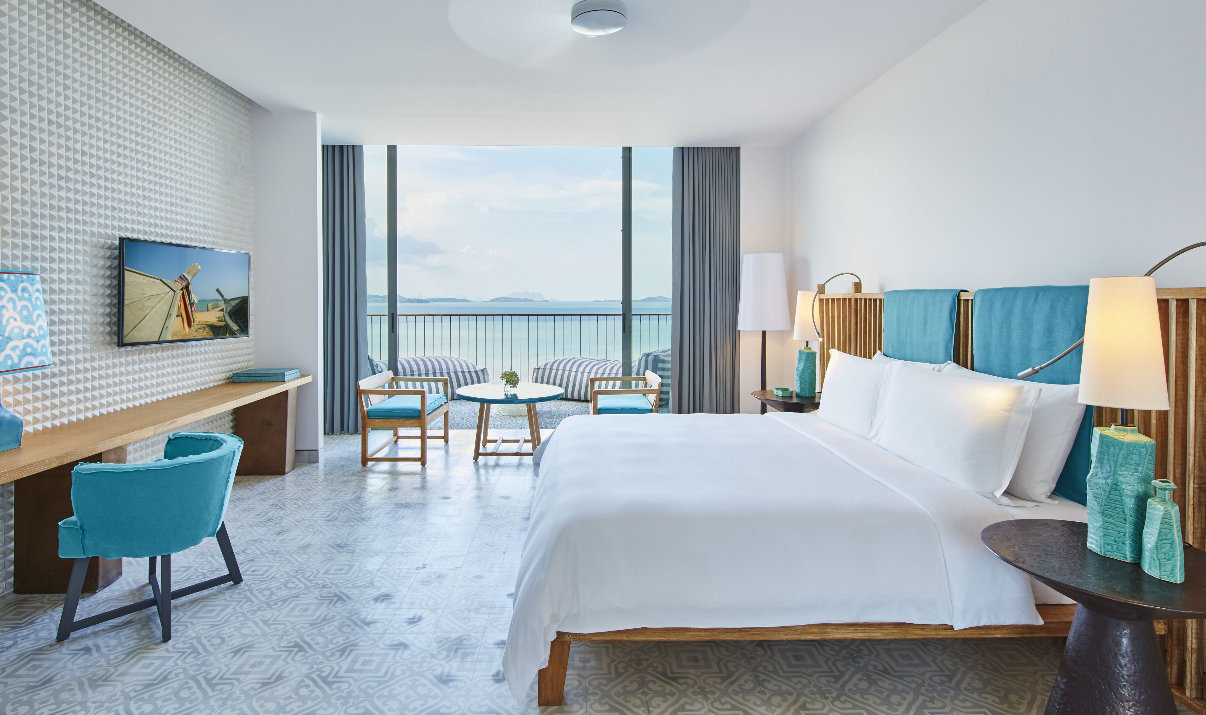 A hotel room with a large bed, a desk, a chair, a small table, and a view of the sea.