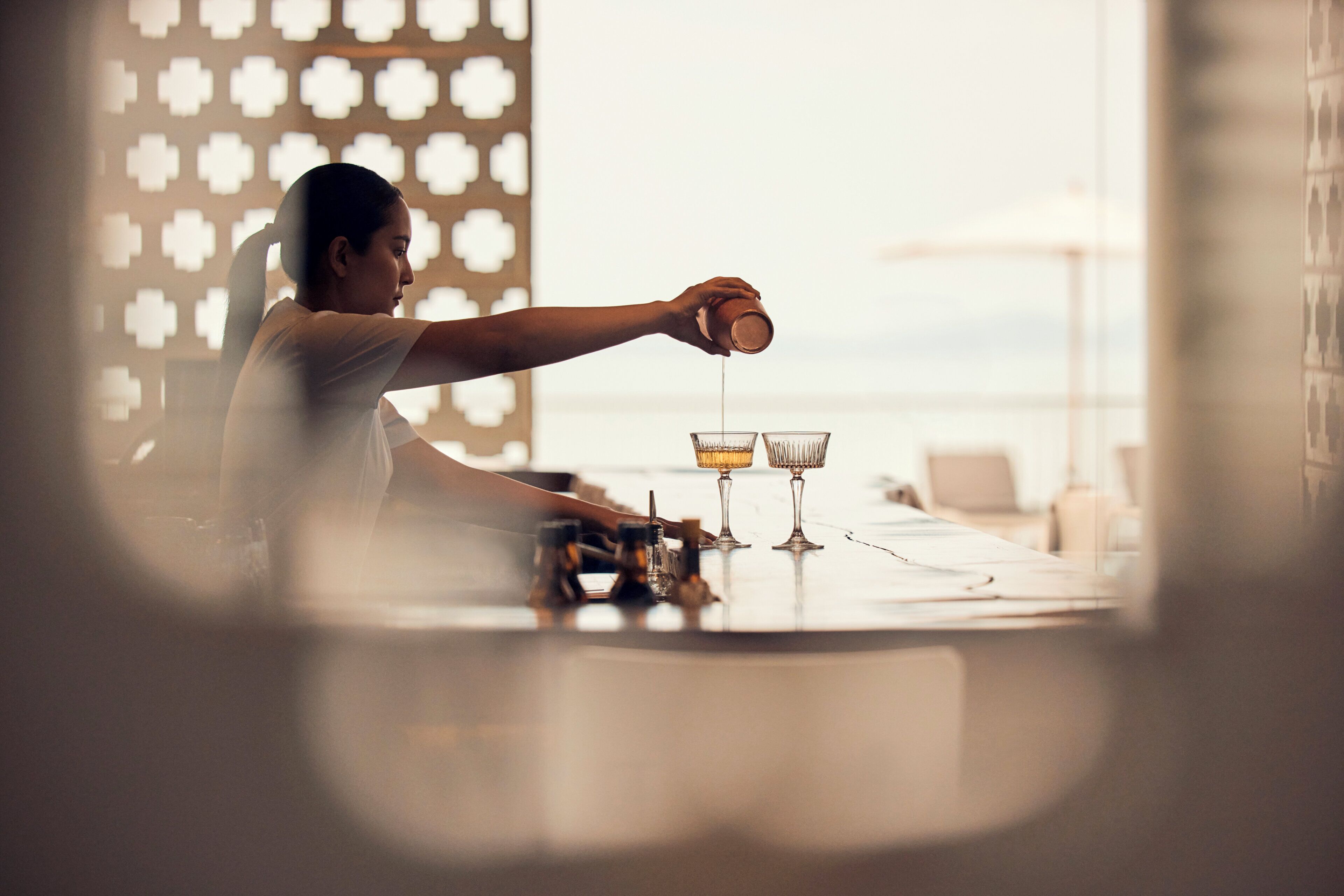 A person pouring a drink into a glass at a bar with a view of the sea.