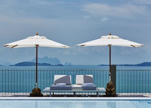 A poolside area with two white lounge chairs under umbrellas, a small statue, and a view of the sea and distant mountains. - COMO Point Yamu, Phuket (Pa Klok)